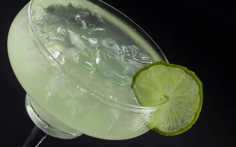 A glass of champagne margarita with a shaker and leaves in a wooden tray