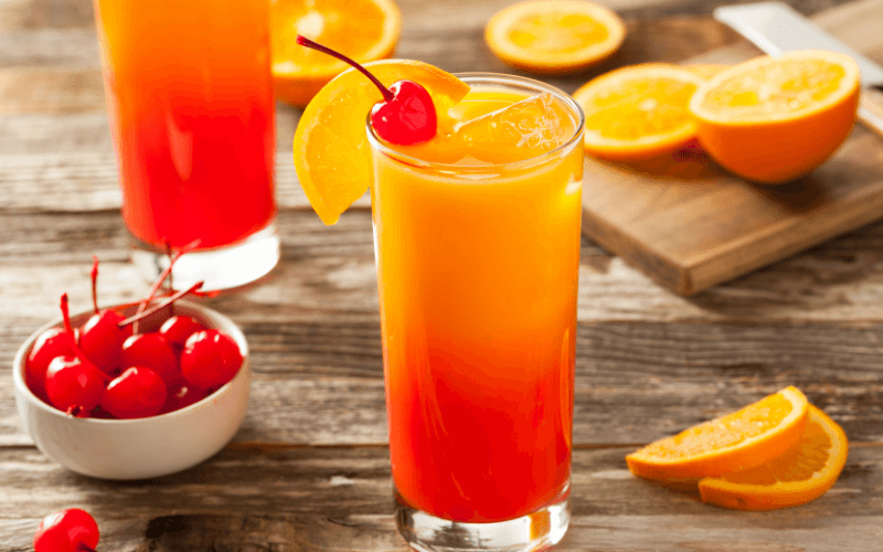 Glasses of tequila sunrise with cherries and oranges on a wooden surface