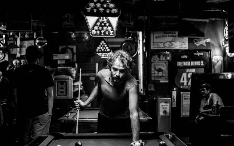 Grayscale photo of a man playing pool in a dive bar