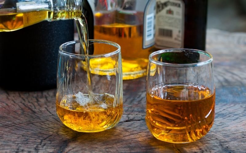 Two glasses with whiskey pouring in over a wooden table