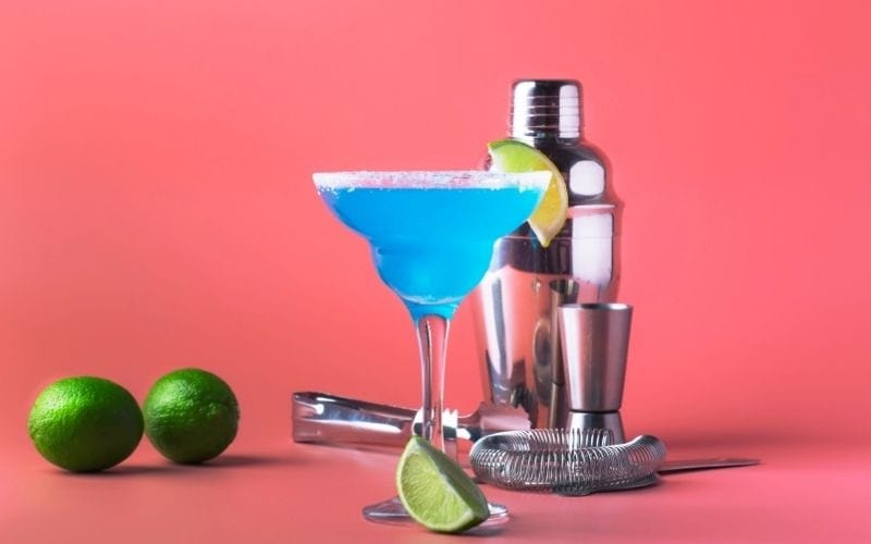 A glass of blue margarita with lime wedges and bartending tools
