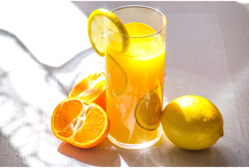 A glass of orange soda garnished with lemon and orange slices surrounded by a lemon and an open orange