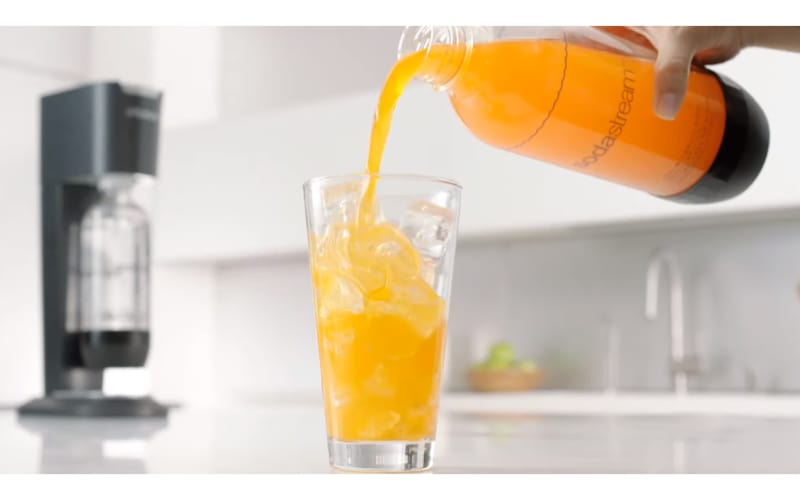 Orange drink being poured into a glass with a SodaStream machine at the back