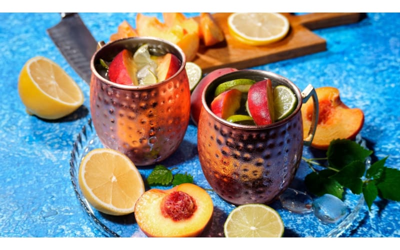 Peach and Ginger Moscow mule served in two copper mugs garnished with lemon and peach slices