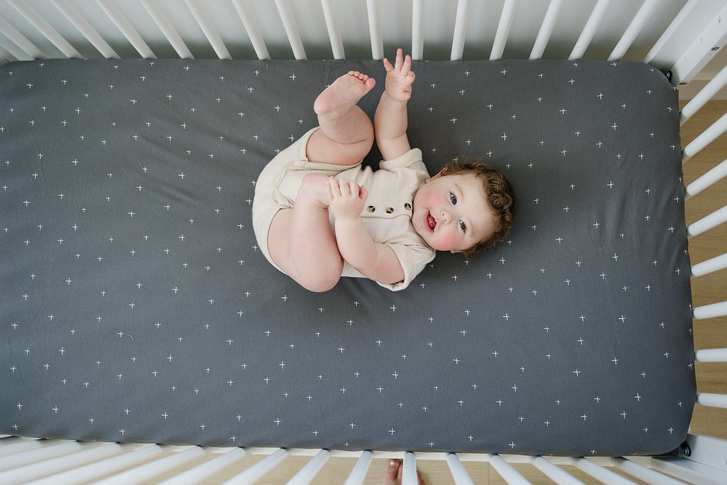 Happy Baby lying on back in crib playing with their feet on an Ecolino Organic Cotton crib sheet in Slate Twinkle