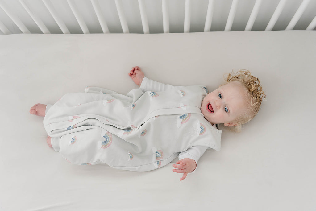 Happy baby lying on back in crib wearing a Woolino 4 Season Sleep Bag with Feet in Rainbow print.