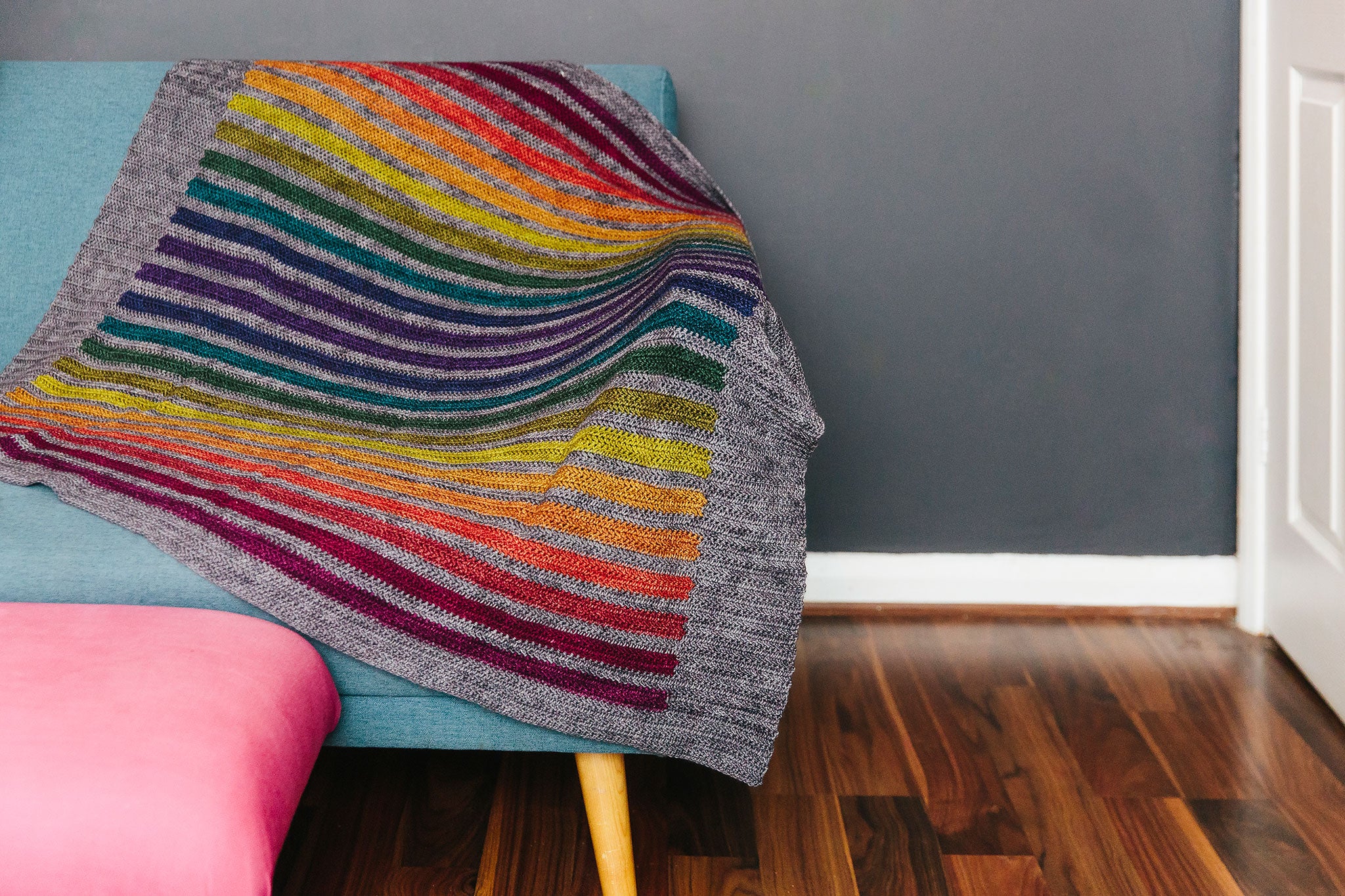 Stormy Rainbow blanket is on a sofa.  12 earthy rainbow stripes crocheted with a dark grey contrast stripe and border.