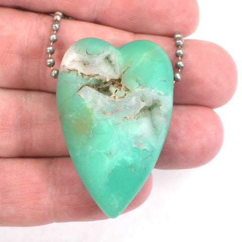 A green chrysoprase heart bead in a person's hand.  The bead has a healed fracture and a vug in it.  