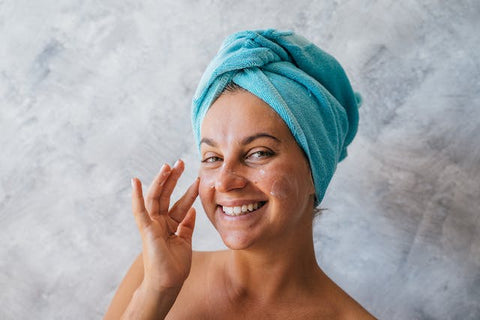 A woman with a towel on her head smiling while using cream on her face
