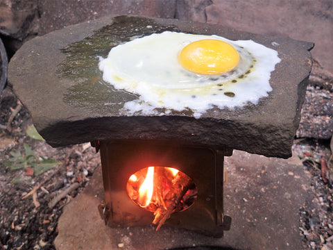 The Überleben Stöker Flatpack Stove is used with a flat rock on top to cook an egg in the wilderness.