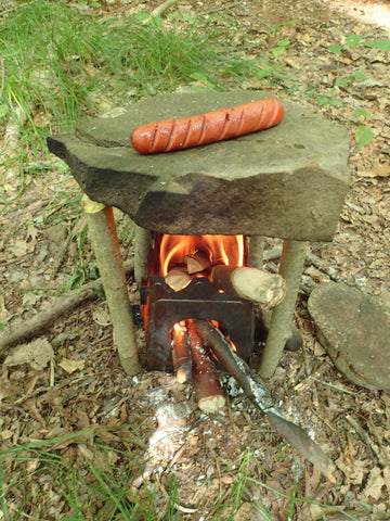 The Stöker Flatpack Stove cooks a sausage with the use of a makeshift griddle stand while in the outdoors.