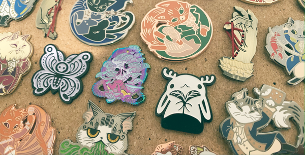 Image of a wide variety of enamel pins designed by missdaisydee. There is a black and white jackalope soft enamel pin, a barfing trinket cat pin, a black and white moth, and a rainbor skull with glowing mushrooms. There are several pins from the Hameowlton Series, Hamilton pins but with cats and puns!