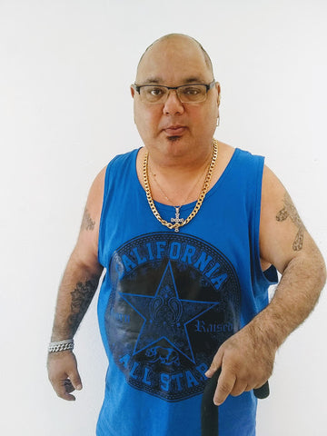 NIAD artist Louie Spagnola, a man with light skin and tattooed arms wearing black frames and a blue tanktop with gold chains around his neck and a black cane.