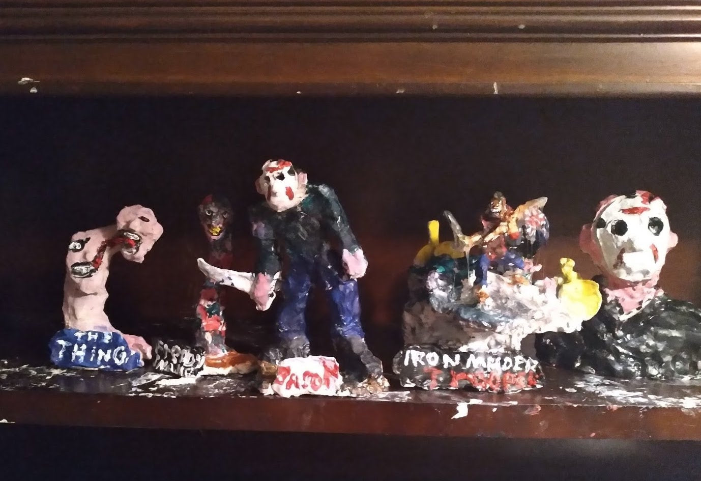 a row of painted mixed-media sculptures lined up on a wooden shelf at the artist's home. Each sculpture depicts a different movie villain or metal band. On the far left is a sculpture of a figure with a long neck and a head that seems to be splitting in two. Text written on the base reads "The Thing." To the right are sculptures of the head and neck of movie villain "Freddie" from Nightmare on Elm street, with his yellow teeth and red and black striped shirt. In the center of the image is a sculpture of movie villain Jason from Friday the 13th: a person wearing a white hockey mask stained with blood, striding forward wielding a machete. To the right of Jason is a sculpture of what appears to be a group of figures on a rocky surface, labeled "Iron Maiden." And finally, to the far right sits a larger bust of Jason, with a black shirt, pink skin, and the same blood stained hockey mask. The shelf is spotted with white paint.