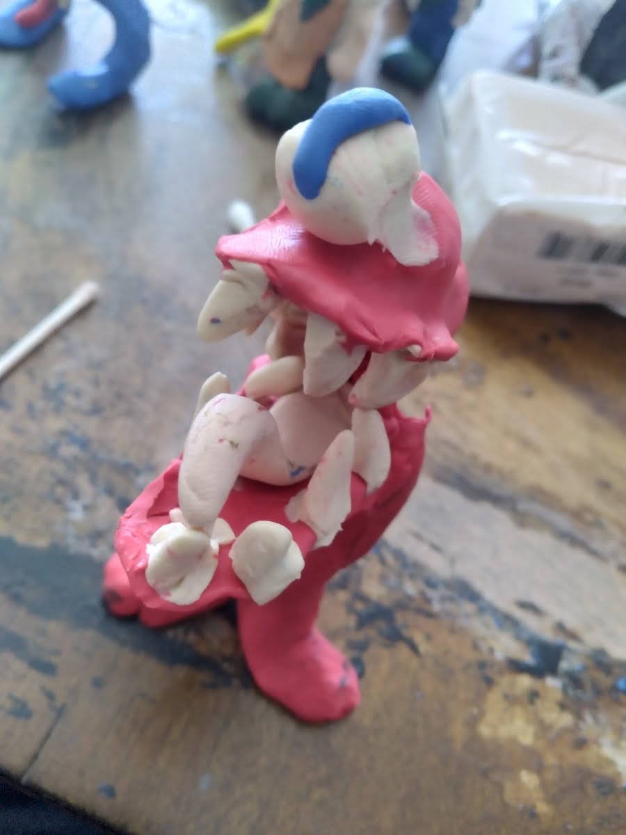 a photograph of a colorful air dry clay sculpture standing on a table. The sculpture depicts a playful and scary red creature: a cyclops with a huge white eyeball with a blue sliver iris, and below a gaping mouth full of sharp white teeth and a curled white tongue. The proportions of the creature appear to be mostly a large head with 2 little red feet sprouting from the  bottom jaw, and stuck to the table to support the sculpture.