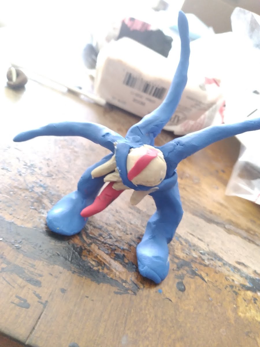 a photograph of a small blue sculpture standing on a wood table. The sculpture is made with air dry clay, and depicts a monster creature shaped like a starfish with spindly radiating arms. The creature has one big white eyeball in the center of its body with a vertical red bar for an iris. Below the eye, sharp white teeth and a long red tongue protrude toward the table. Three of the creature's limbs stretch out and up around its head, and two are bent and planted on the table, keeping the character standing upright.