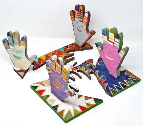 Four wooden hands, positioned upright on the square panel from which they were cut, decorated in horizontal bands of vibrant colors on each finger and jagged patterns along the edges of the base, a smiling face on each hand.