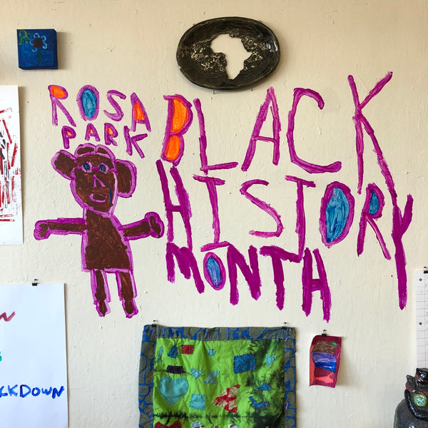 A detail image of a portion of the gallery wall. There is a painting of a figure colored brown and purple, with the words "Rosa Parks Black History Month" written in purple around them. Above this wall painting, there is a ceramic plate with a cut out in the shape of the African continent installed on the wall. To the right of this plate there is a small square blue painting of a flower. Along the bottom of this image, there is a green and blue quilted textile with hearts and other shapes appliquéd on it. To the right of this textile piece there is a small maroon and pink abstract marker drawing. The edges of the photo show corners of additional drawings that are installed on the wall, beyond the scope of this photograph.