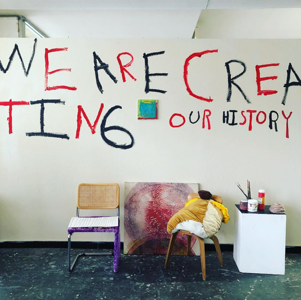 this is a photograph taken early on in the set up of an exhibition, and features a wall mural, some paintings and sculptures that have not yet been installed. There is a white wall with the words "WE ARE CREATING OUR HISTORY" painted in black and red capital letters. There is a small blue and yellow and red square painting hanging in the center of the wall. A large painting of a red and purple circle sits on the floor and leans against the wall at the center of this photo. To the left of this painting there is a tan and purple chair; to the right and in front of the painting there is a brown chair with a stuffed pillow sculpture sitting on it. There is a white pedestal to the far right of this image that has paint tubs and brushes on it. The floor in front of the wall is dark mottled grey.