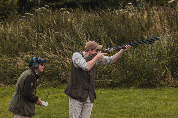 A shooter takes aim during a clay shooting session with a ShotKam Gen 4 Mini attached to the barrel, while an instructor assists.