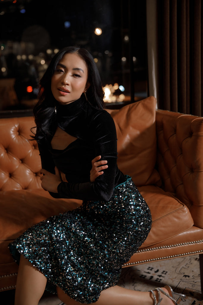 Model sitting with arms folded wearing Black Stretch Velvet Bodysuit and Emerald Sequin Skirt