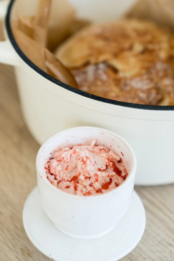 Close up photo of homemade strawberry butter in round butter dish with homemade dutch oven bread in the background