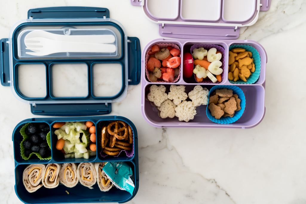 Overhead image of 2 bento lunch boxes- one navy blue (left) and one lilac purple (right) with pinwheel style sandwiches, peanut butter and jelly sandwiches cut in the shapes of flowers, pretzels, goldfish, and various fruits and veggies cut into heart, star, and flower shapes