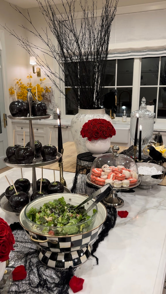 Image of Rachel Parcell's kitchen countertop filled with black Halloween decor, a vase of red roses, and food including a salad, black caramel apples, and pink macaroons with vampire fangs painted on