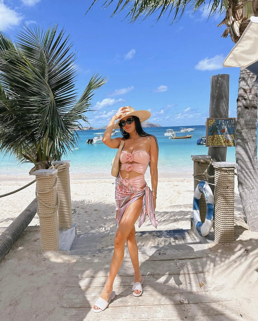 Rachel Parcell standing on beach in St. Barths wearing pink floral detailed one-piece swimsuit, pink sarong, sunhat and sunglasses