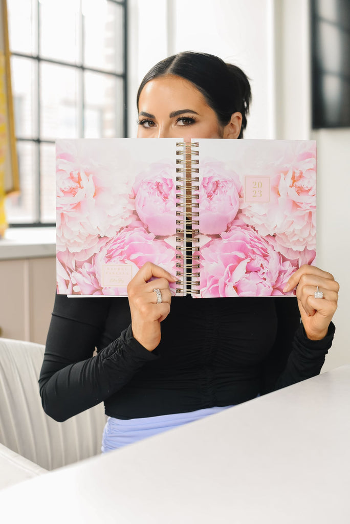 Rachel Parcell hiding face with RP x Blue Sky 2023 Planner with pink peonies and looking at camera 