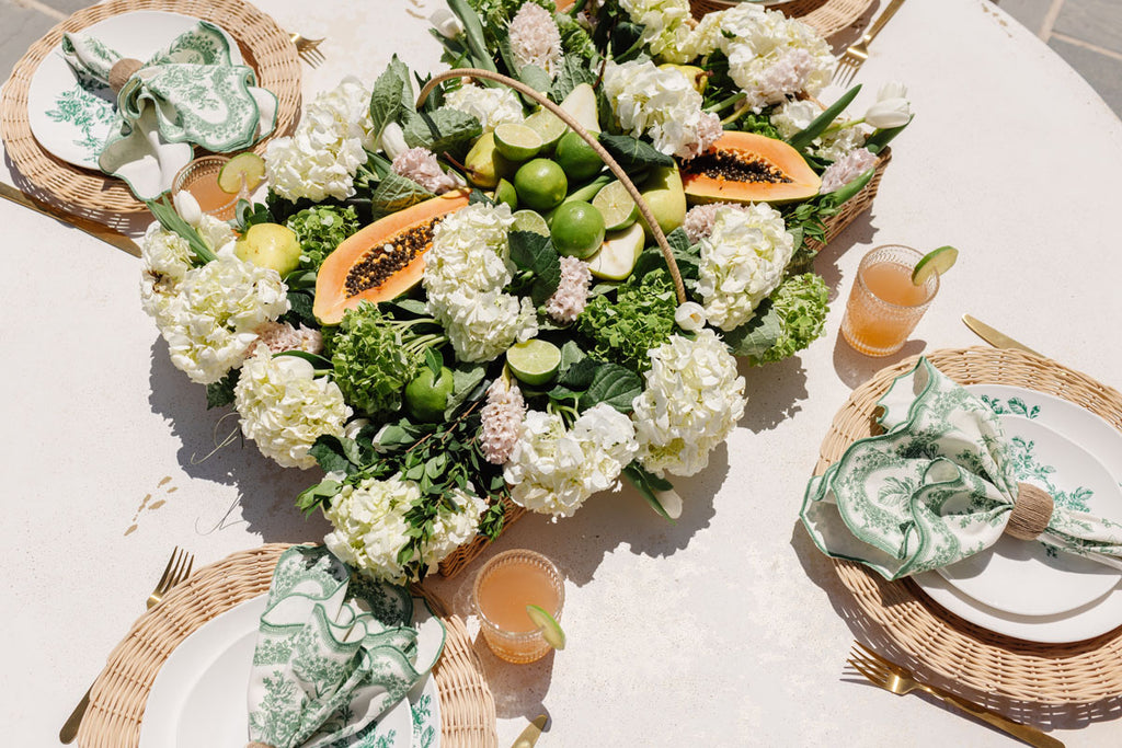 Rachel Parcell Summer 2022 Home collection table setting with RP Wicker Basket center piece filled with white hydrangeas, white tulips, limes, and papayas 