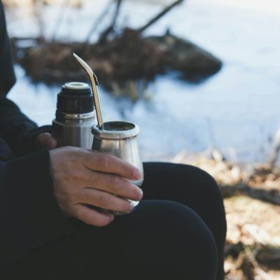 A person resting a gourd of Yerba Mate with a thermos filled with warm water outdoors.