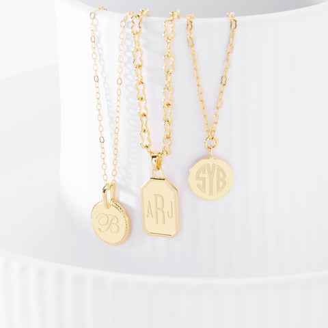 Three gold necklaces dangle, showing initial and monogram engravings on their pendants.