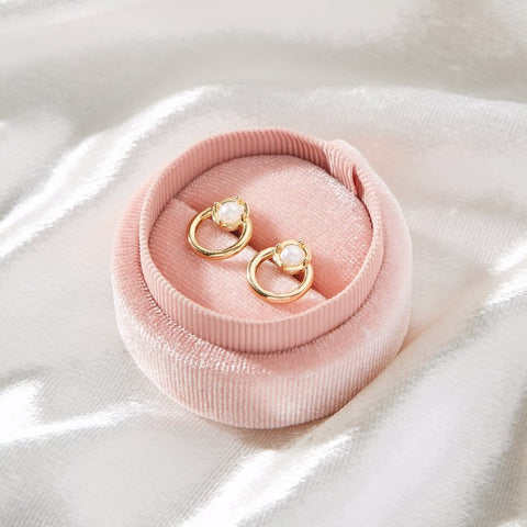 A set of gold stud earrings with pearl inlay are nestled into a round, pink jewelry box.