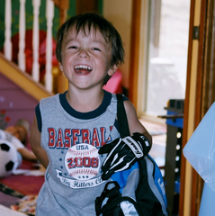 Michael Zeiter at age three laughing at the camera and holding his t-ball baseball gear in his family home at Our Lady's Ranch in Grass Valley, CA.
