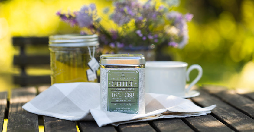 A tabletop outdoors with flowers, tea brewing in a mason jar and a tea cup all in the background. In the front is a tin of Chamomile mint CBD tea from Harney & Sons