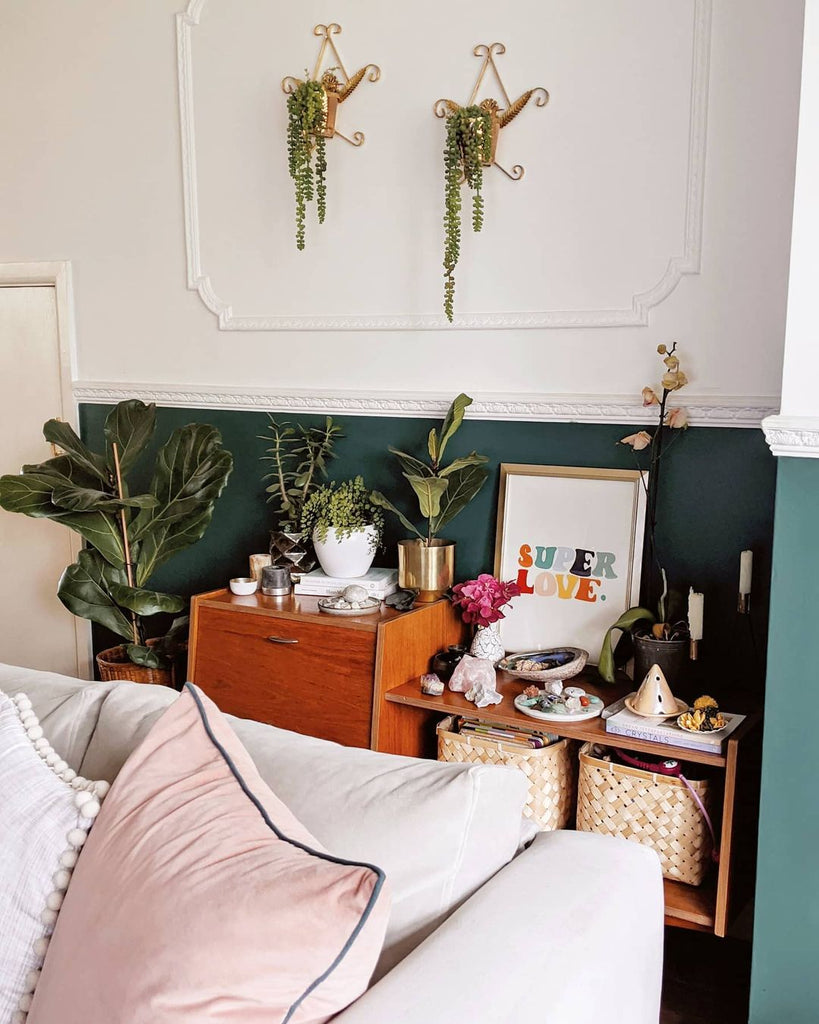 bright, sunny living room painted half-height in a deep green colour and filled with plants, vintage furniture and a rainbow print nestled on the side cabinet