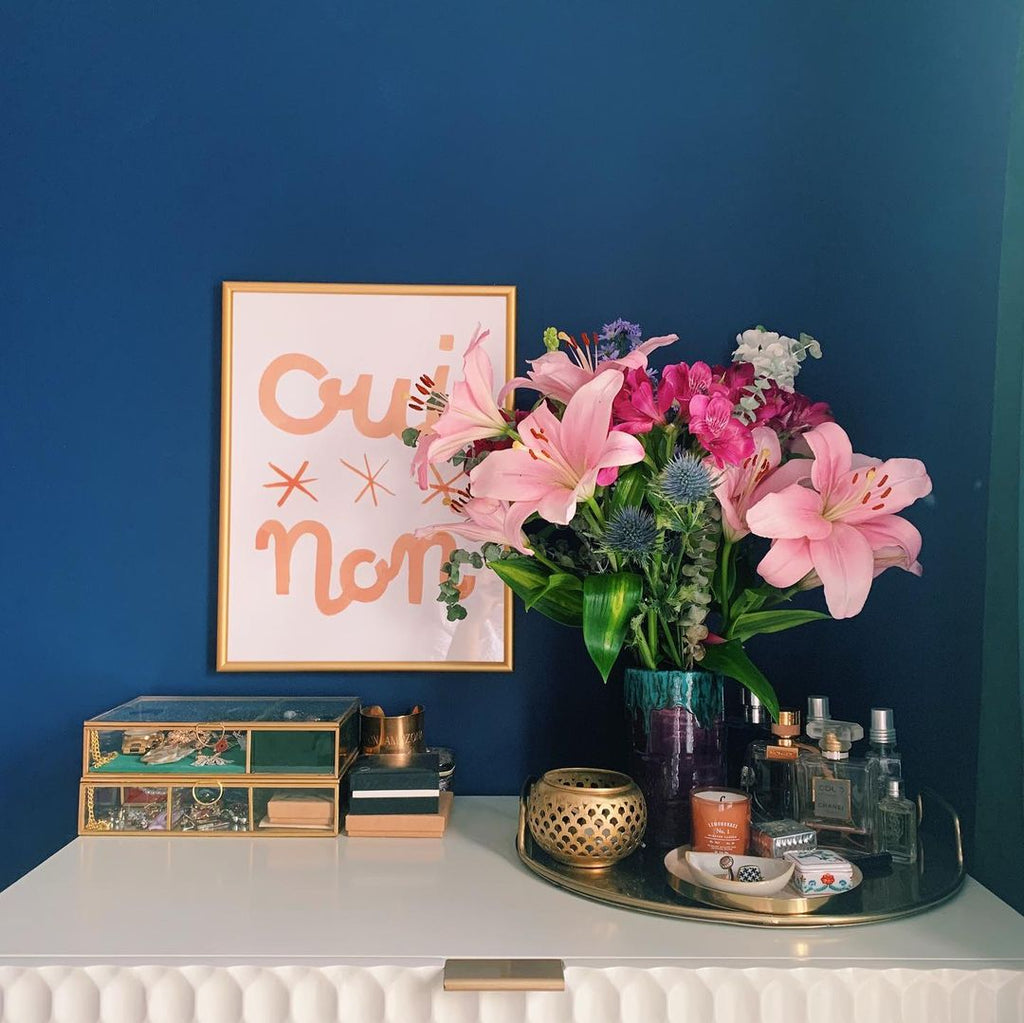 A bedroom painted deep blue, with a white textured dresser. On top stands a bunch of pink lilies, jewellery storage, and above it a pink lettered french print hangs on the wall