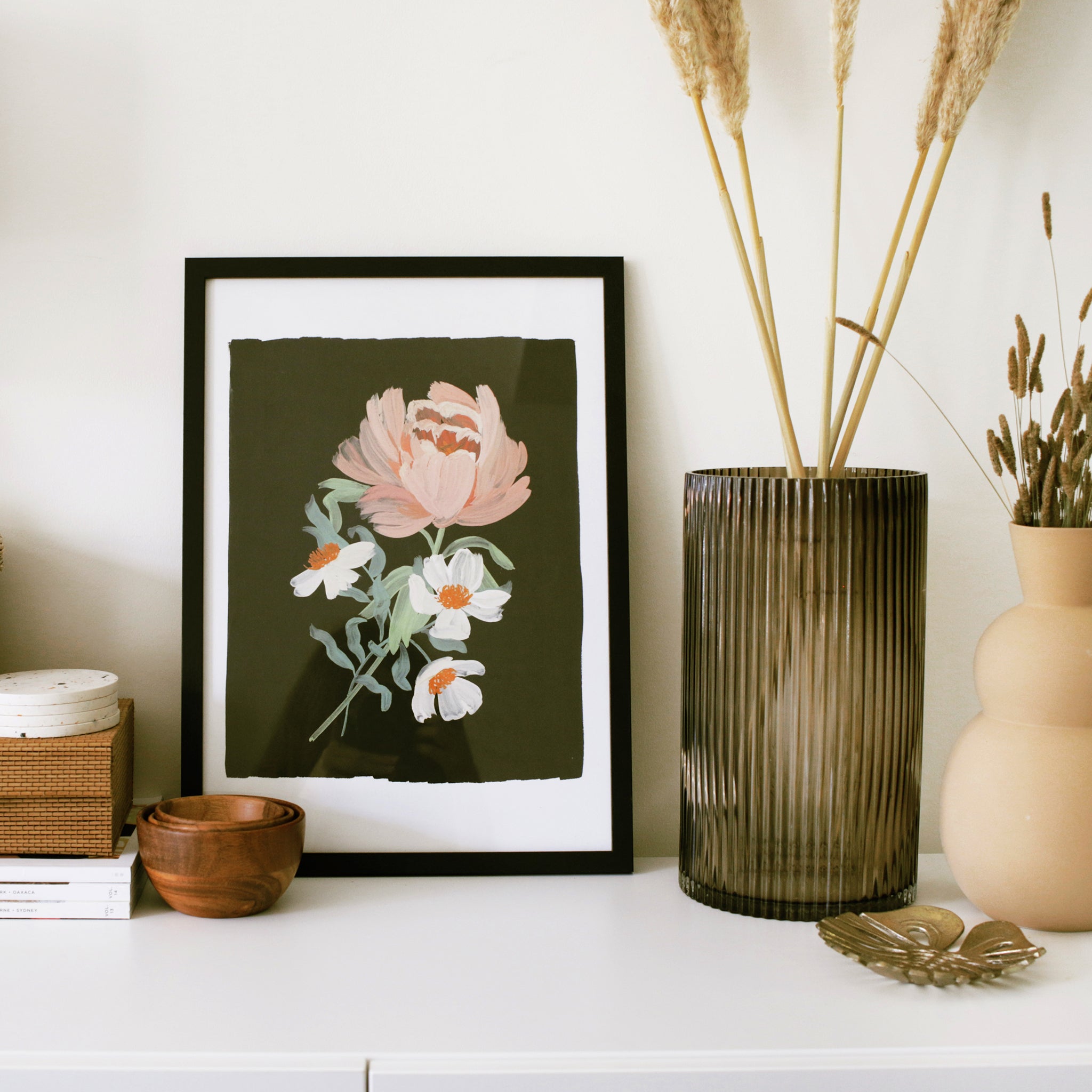 a black floral print featuring a beautiful pink peony sits in a black frame, on top of a white cabinet, decorated with minimal, natural vases and trinket boxes