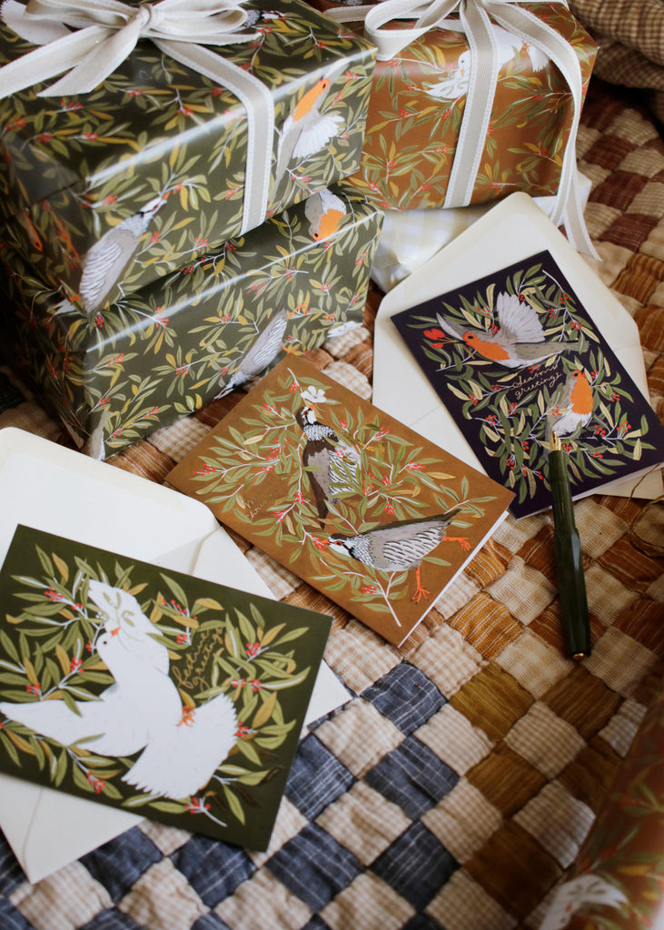 a set of three christmas cards featuring christmassy birds (robins, doves and partridges) lay next to a stack of gifts wrapped in matching bird-patterned wrapping paper