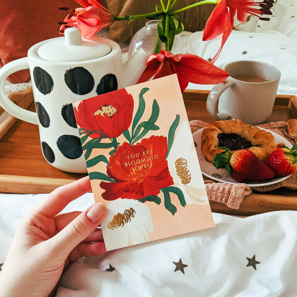 Holding a floral card that reads "you are blooming lovely" infront of a breakfeast-in-bed tray