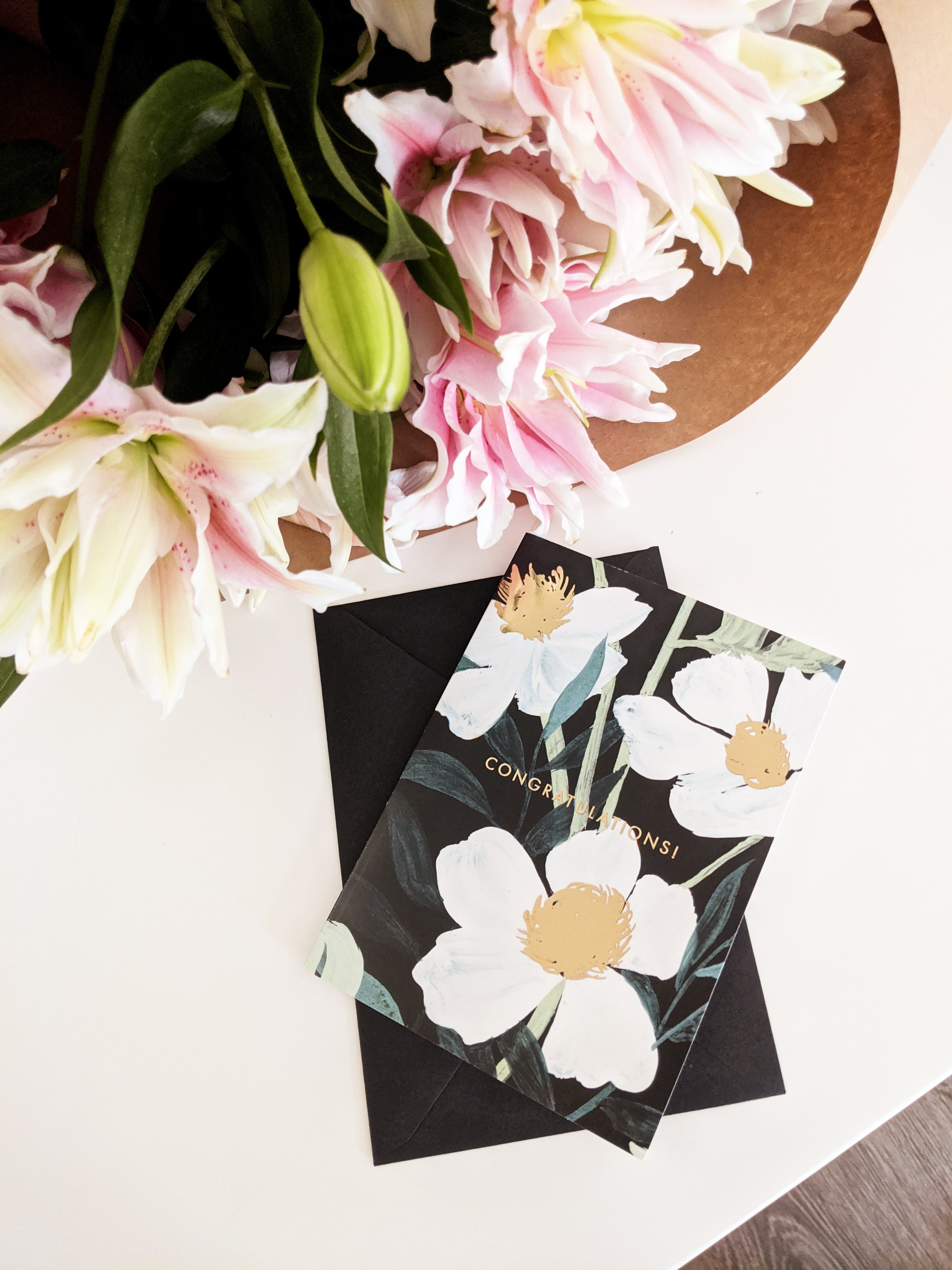 An elegant, black floral Congratulations card sits on a table alongside a bunch of gorgeous pink lilies