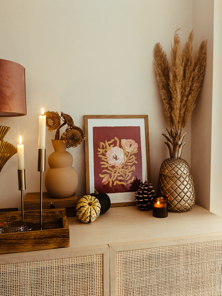 a brown floral print leant on a rattan sideboard, surrounded by dried flowers, candles and decorative pumpkins