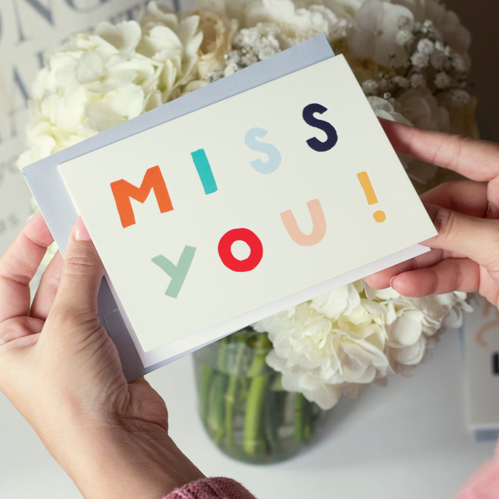 holding a pale yellow card that reads "MISS YOU!" in bold, rainbow letters. 