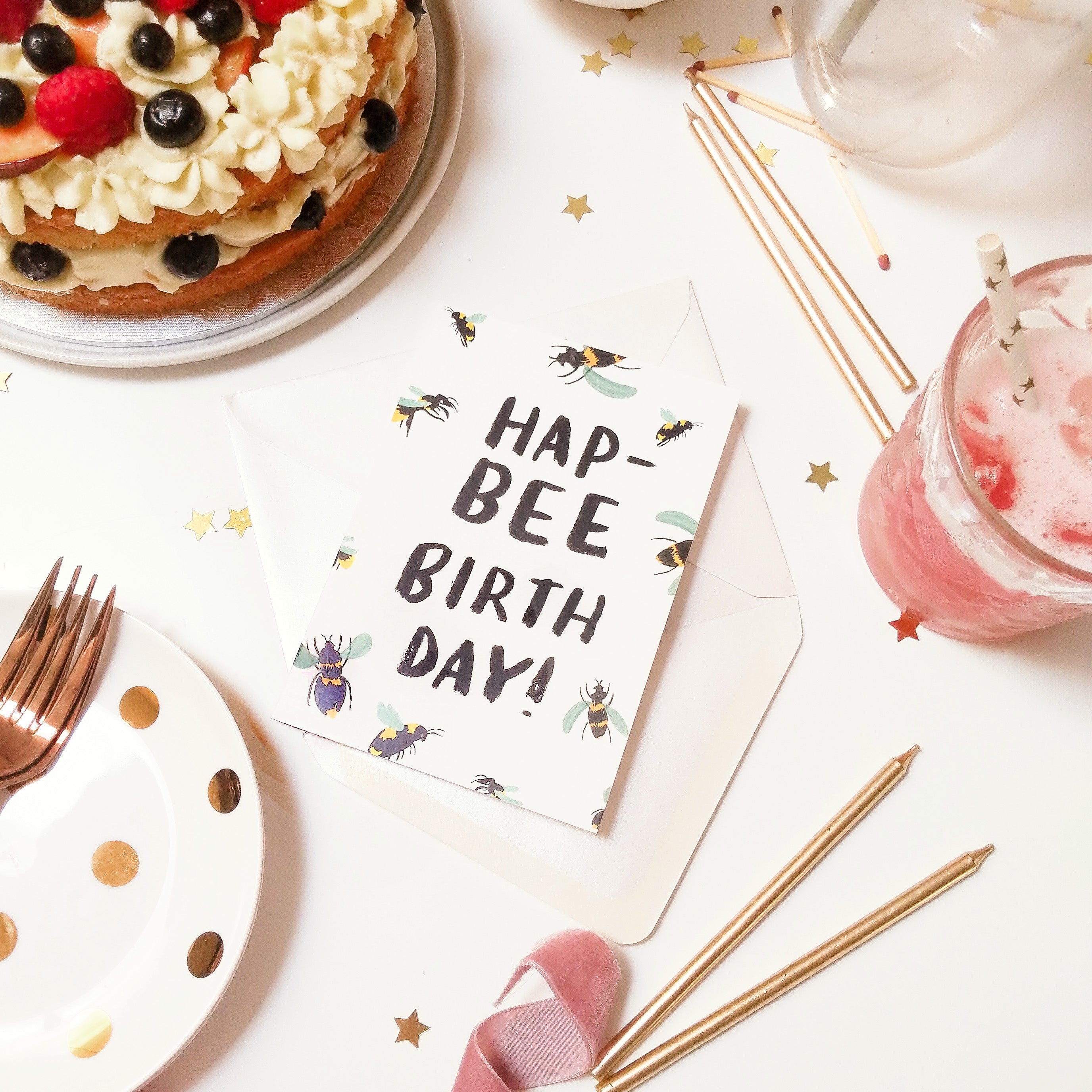 The hapbee birthday card lays on a table surrounded by cake, candles and ribbon