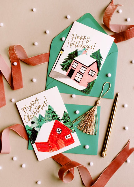 two christmas cards, illustrated with snow covered buildings with teal envelopes on a grey background, surrounded by pink velvet ribbons and mini white pompoms 