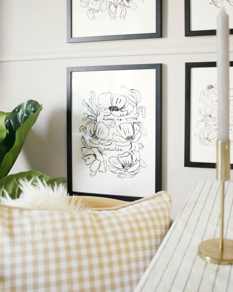 an illustrated floral print hangs in a black frame against a pale grey wall - it reads "home is paradise" surrounded by black line art flowers 