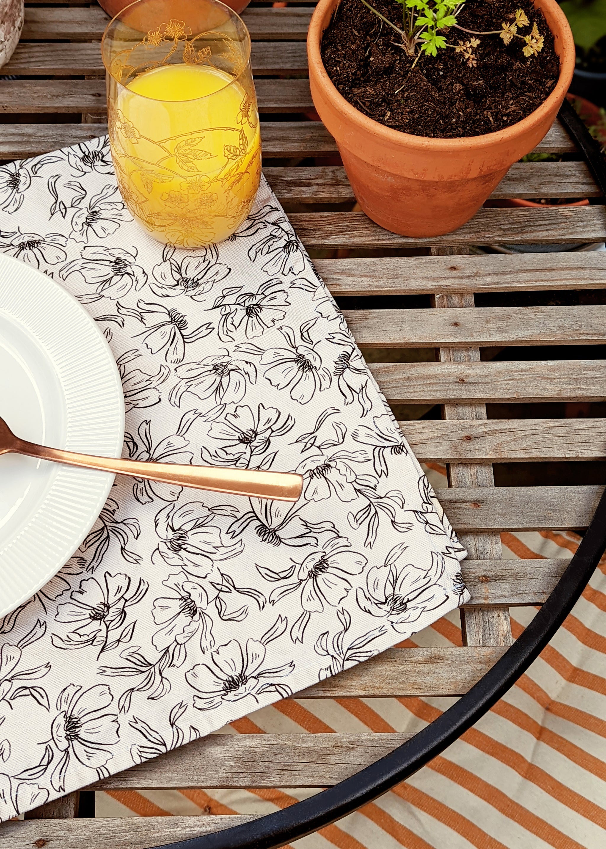 a black and cream, linework-style floral patterned tea towel spread out on a wooden garden table, used as a place setting / place mat