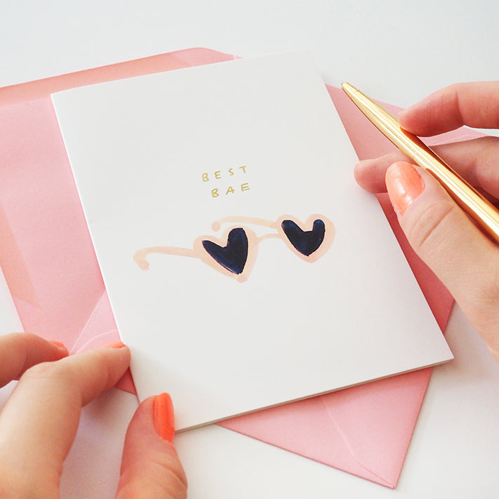 holding a gold pen, about to write inside a white valentine's card that reads "best bae" in gold, above a pair of illustrated heart shaped sunglasses.
