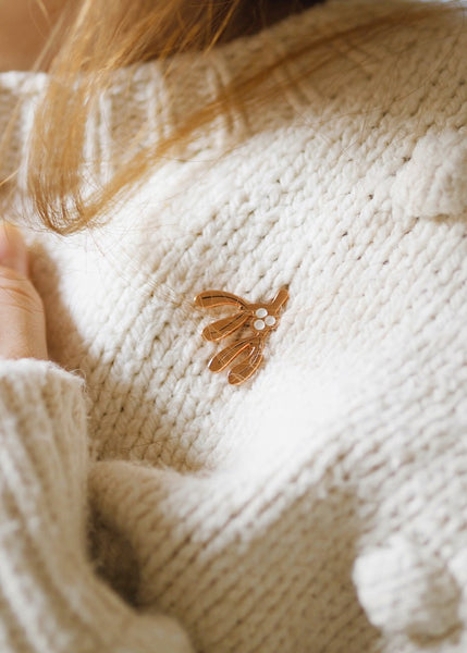 A rose gold enamel pin in the shape of a sprig of Mistletoe, pinned on a soft white jumper